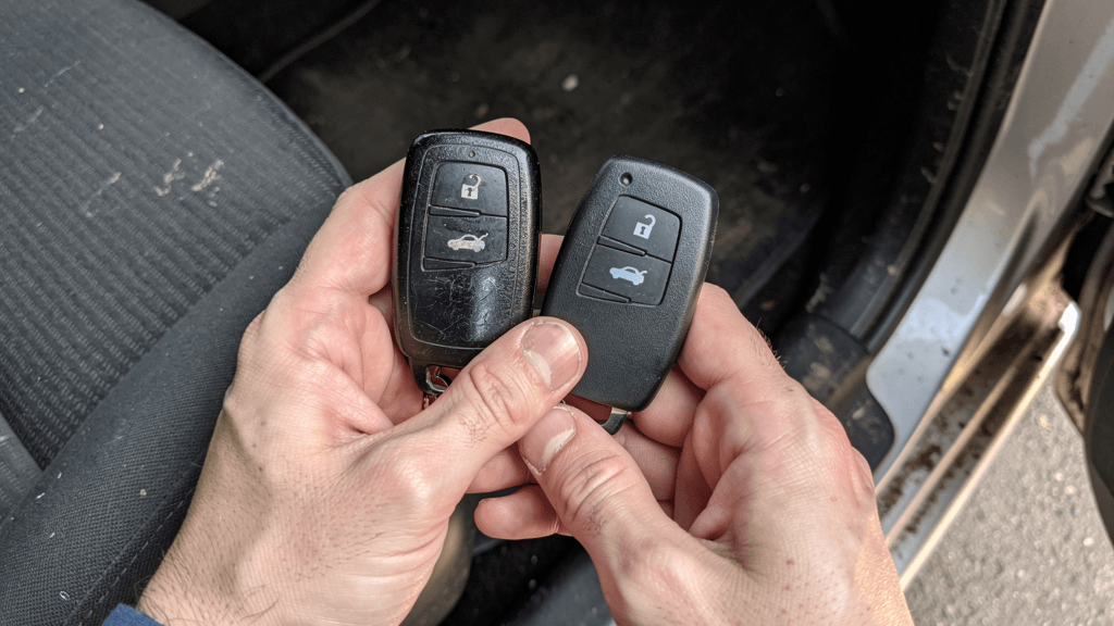 Locksmith comparing a worn key fob with an unbranded replacement beside a car